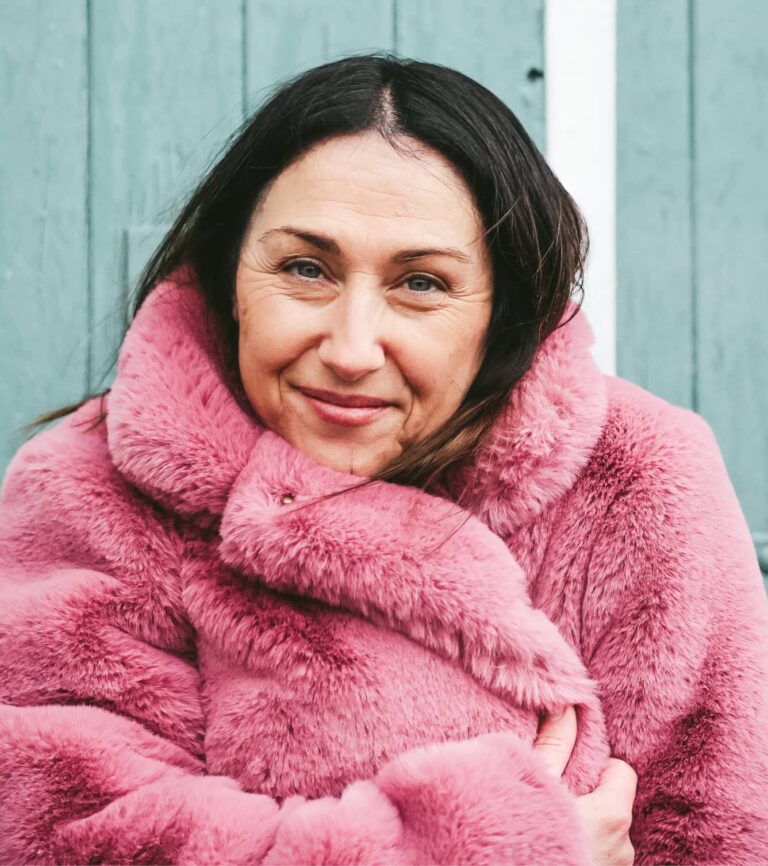 Khalie smiling warmly while wearing a pink faux fur coat against a teal background.