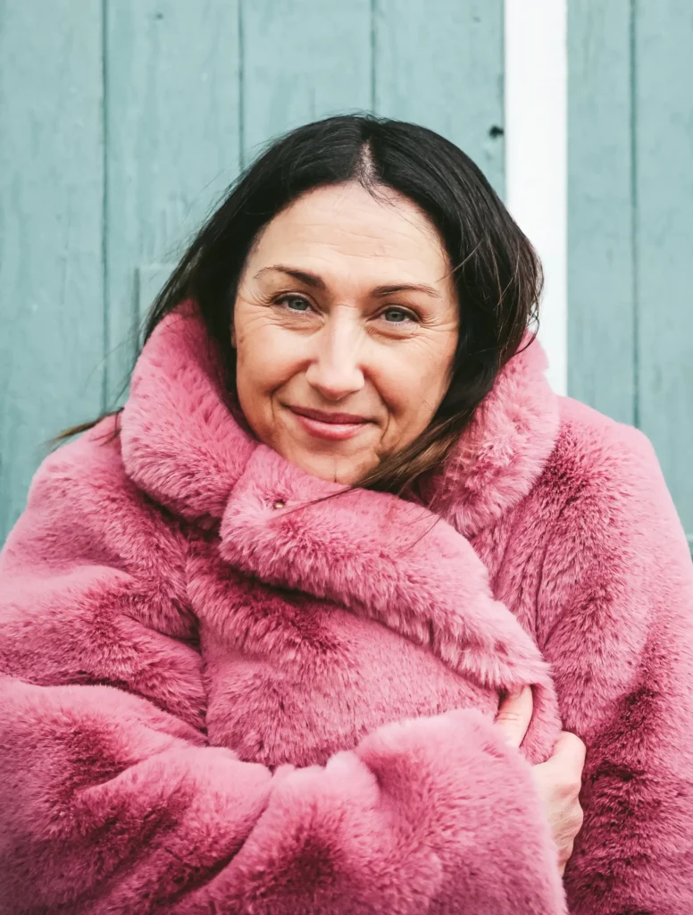 Khalie smiling warmly while wearing a pink faux fur coat against a teal background.